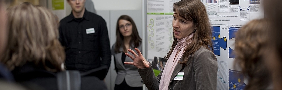 Konferenzimpression: Eine junge Frau erklärt gerade einer um sie versammelten Gruppe ein wissenschaftliches Poster